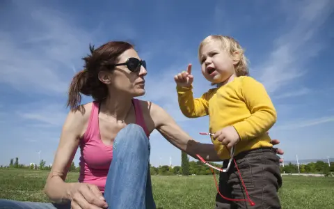 Niña rubia con camiseta amarilla, con el dedo levantado, parece que está aprendiendo cuando empiezan a balbucear los bebes. Madre con gafas de sol la mira.