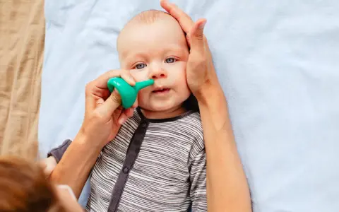Bebé con ojos azules siendo ayudado a quitar mocos con un aspirador nasal verde.