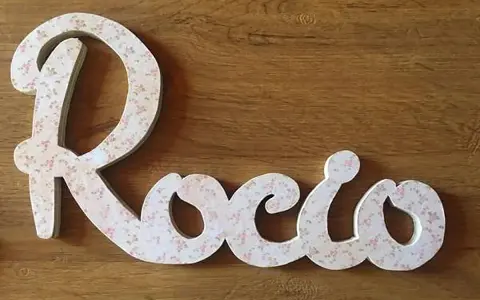 Letra decorativa con el nombre Rocio, cubierta de un delicado estampado floral rosa sobre fondo blanco, sobre una superficie de madera.