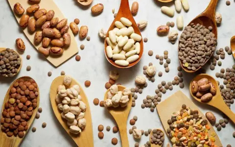 Cucharas de madera repletas de legumbres bebes: lentejas, garbanzos y frijoles de diversos tipos, esparcidos sobre una superficie clara.