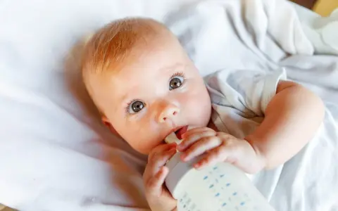 Bebé con ojos grandes y curiosos, bebiendo leche de un biberón. La imagen responde a la pregunta: cuanta leche toma un bebe.