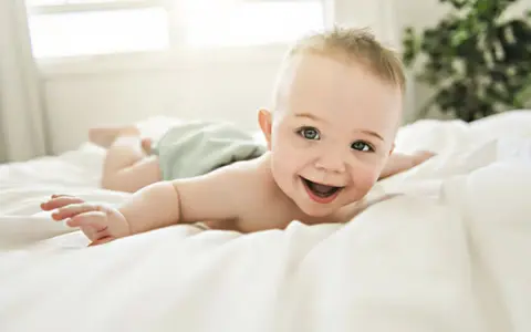 Bebé sonriente boca abajo en una cama blanca, con un reflejo de hociqueo en su expresión juguetona.