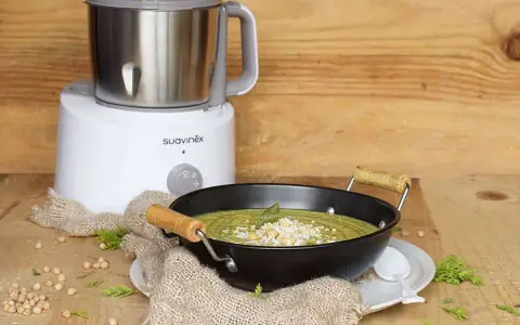 Un puré verde cremoso, ideal para nabo bebés, servido en un cuenco negro con garbanzos y hierbas. Al fondo, una máquina de cocina Suavinex.