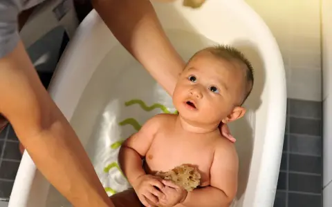 Bebé en bañera, manos adultas ajustan la temperatura del agua. Sostiene una esponja, mirando hacia arriba con curiosidad.