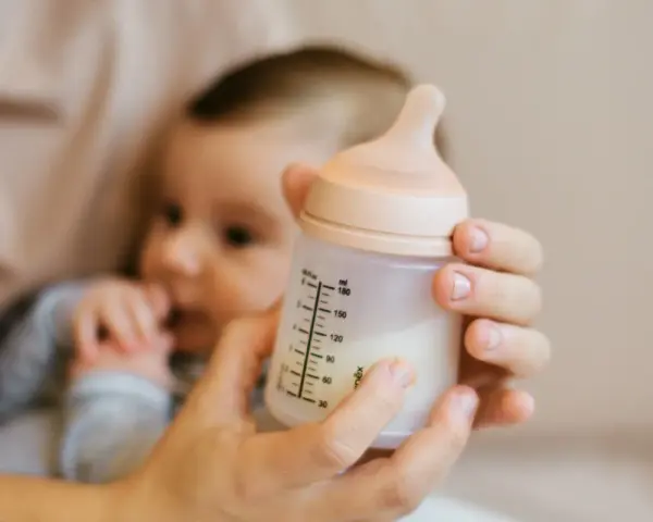 Manos sostienen un biberón con leche, con un bebé borroso al fondo. Es hora de considerar cuando cambiar tetina biberon.