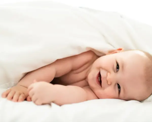 Un bebé sonriente se asoma bajo una manta blanca. Parece que los bebes tienen memoria, ¡y este ya recuerda cómo sonreír!