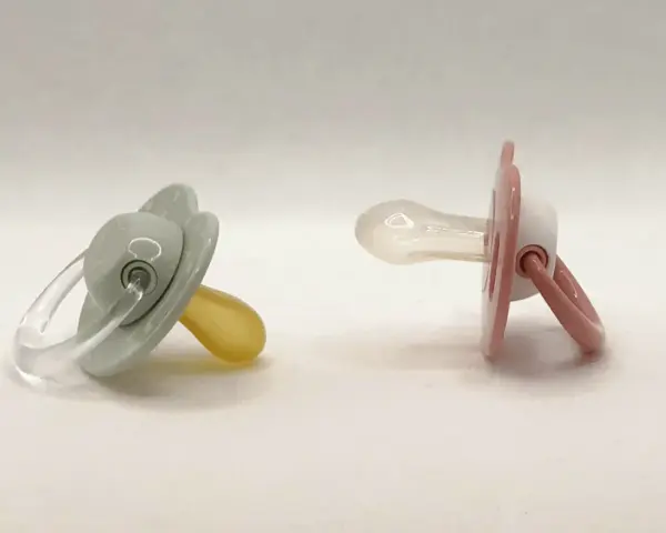 Dos chupetes, uno gris con tetina de látex y otro rosa con tetina de silicona, esperan a ser elegidos.