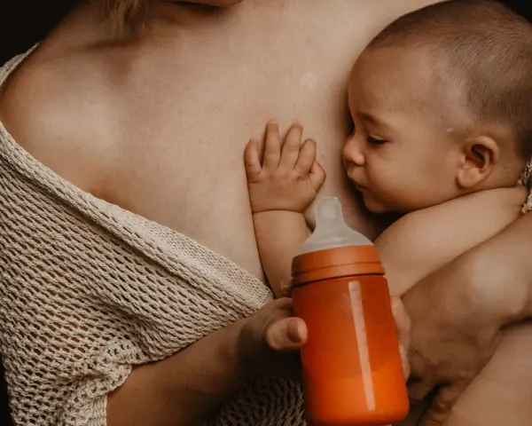 Bebé succionando un biberón naranja, con la mano apoyada en el pecho de la madre. Se aprecian varios tipos de tetinas en el mercado.