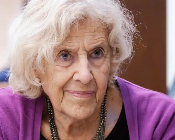 Retrato de una anciana con cabello blanco y ojos azules, luciendo un suéter morado. Su rostro arrugado evoca la sabiduría y el significado de una vida plena, como el nombre Manuela.