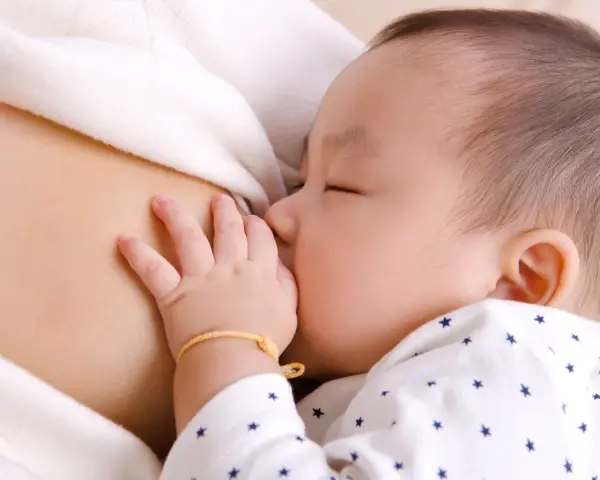 Bebé asiático durmiendo mientras se alimenta, mostrando el reflejo de succión. Su mano pequeña descansa sobre el pecho de su madre.