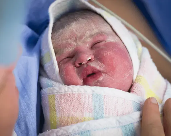 Recién nacido envuelto en una manta, con la piel rosada y cubierta de vernix bebe. Una mano lo acaricia suavemente.