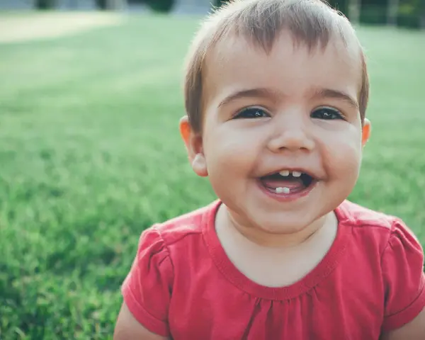 Un bebé de 1 año sin dientes ríe a carcajadas en el césped, mostrando dos pequeños dientes inferiores.