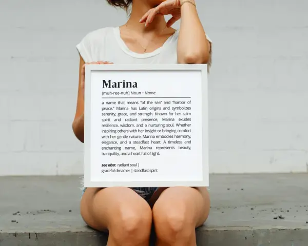 Una mujer sostiene un marco blanco con la definición del nombre Marina, que significa "del mar" y "puerto de paz".