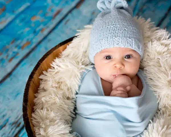 Bebé Ezra, con gorrito azul y envuelto en mantita, mira curioso. Descansa en una cesta sobre fondo de madera azul.