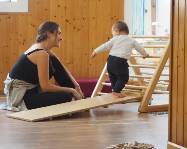 Niña gateando en rampa de madera, animada por mujer sonriente. Fomentando el movimiento libre y la exploración.