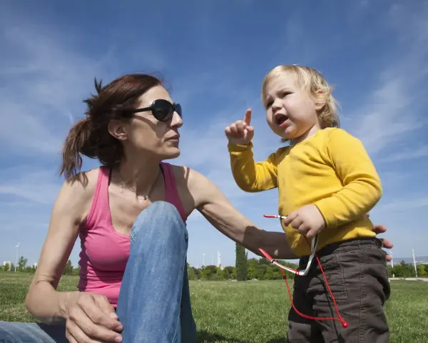 Niña rubia con camiseta amarilla, con el dedo levantado, parece que está aprendiendo cuando empiezan a balbucear los bebes. Madre con gafas de sol la mira.