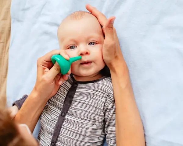 Bebé con ojos azules siendo ayudado a quitar mocos con un aspirador nasal verde.