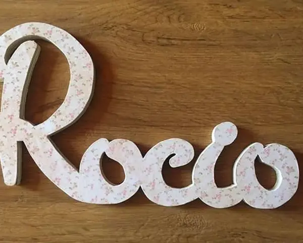 Letra decorativa con el nombre Rocio, cubierta de un delicado estampado floral rosa sobre fondo blanco, sobre una superficie de madera.