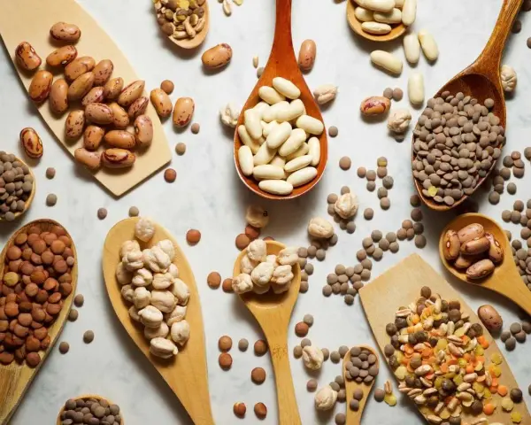 Cucharas de madera repletas de legumbres bebes: lentejas, garbanzos y frijoles de diversos tipos, esparcidos sobre una superficie clara.