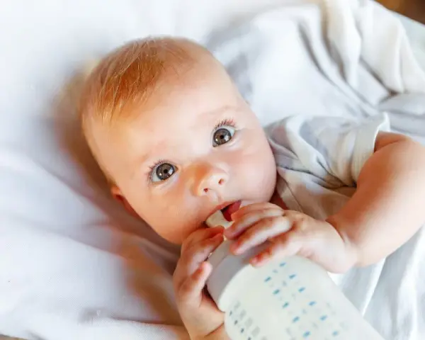 Bebé con ojos grandes y curiosos, bebiendo leche de un biberón. La imagen responde a la pregunta: cuanta leche toma un bebe.