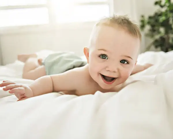 Bebé sonriente boca abajo en una cama blanca, con un reflejo de hociqueo en su expresión juguetona.