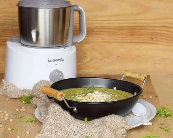 Un puré verde cremoso, ideal para nabo bebés, servido en un cuenco negro con garbanzos y hierbas. Al fondo, una máquina de cocina Suavinex.