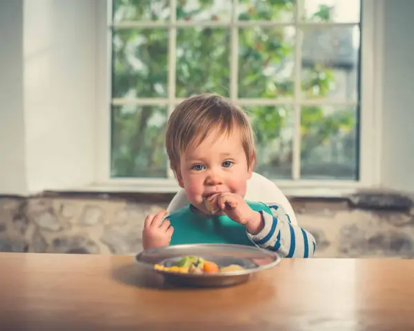 Un bebé con babero verde come felizmente de un plato. Parece que está disfrutando de su comida, quizás marisco para bebés.
