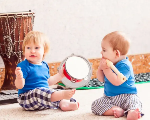 Dos bebés disfrutan de la estimulación musical temprana, uno con un tambor y otro con una flauta, rodeados de instrumentos.