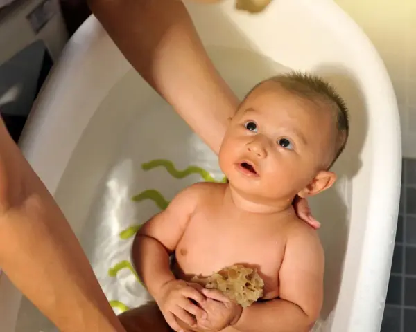 Bebé en bañera, manos adultas ajustan la temperatura del agua. Sostiene una esponja, mirando hacia arriba con curiosidad.