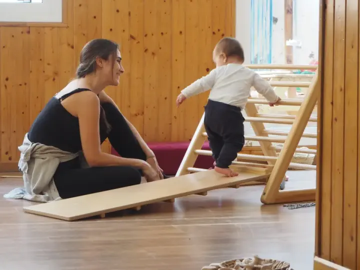 Niña gateando en rampa de madera, animada por mujer sonriente. Fomentando el movimiento libre y la exploración.