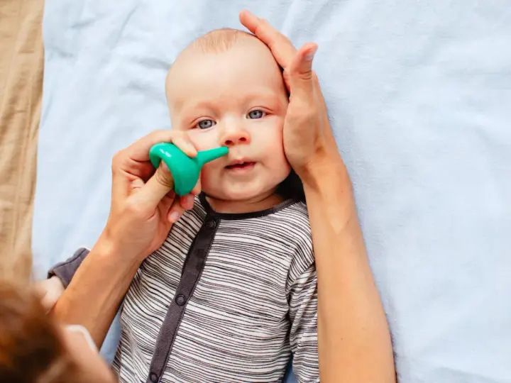 Bebé con ojos azules siendo ayudado a quitar mocos con un aspirador nasal verde.