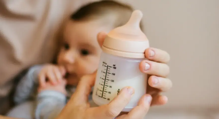Manos sostienen un biberón con leche, con un bebé borroso al fondo. Es hora de considerar cuando cambiar tetina biberon.