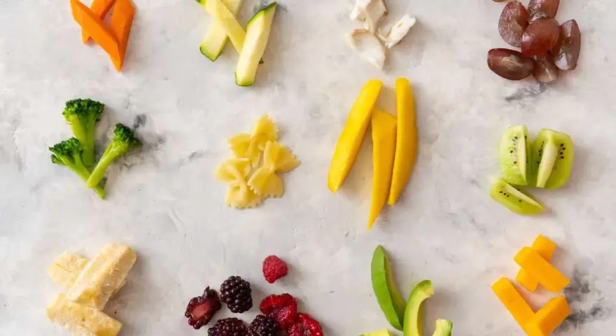 Variedad de alimentos para blw bebé 6 meses: zanahoria, calabacín, brócoli, pasta, mango, kiwi, aguacate, bayas, plátano y queso.