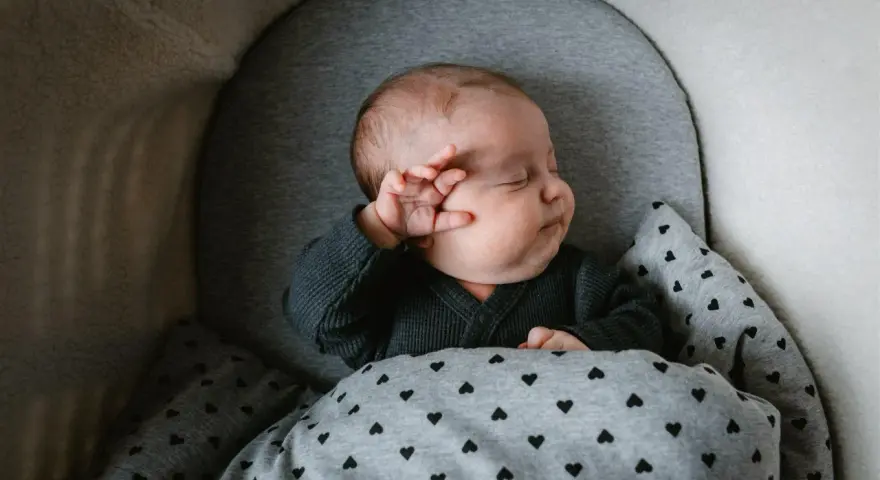 Bebé durmiendo abrigado con un mono gris y manta de corazones. Ideas para como vestir a un recien nacido en junio.