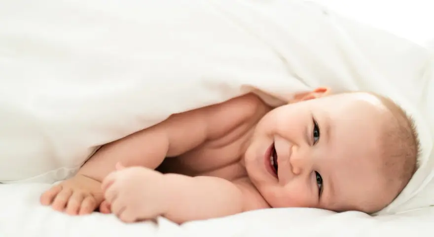 Un bebé sonriente se asoma bajo una manta blanca. Parece que los bebes tienen memoria, ¡y este ya recuerda cómo sonreír!