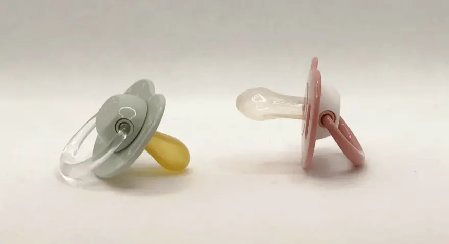Dos chupetes, uno gris con tetina de látex y otro rosa con tetina de silicona, esperan a ser elegidos.