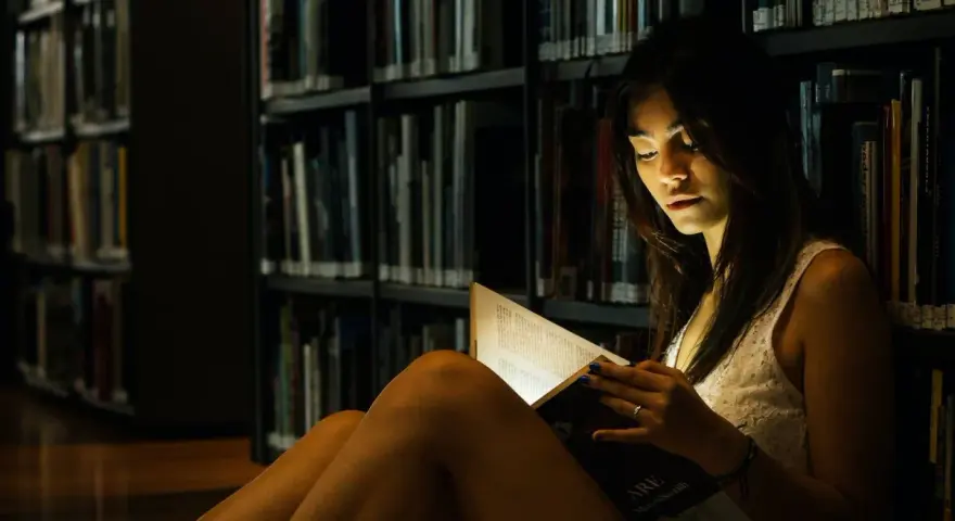 Elena, in the library's dim light, engrossed in a book.
