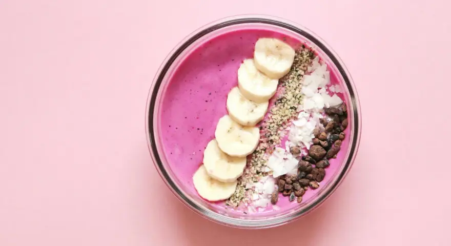Un tazón de batido rosa vibrante con rodajas de plátano, coco rallado, semillas de chía y trozos de cacao.