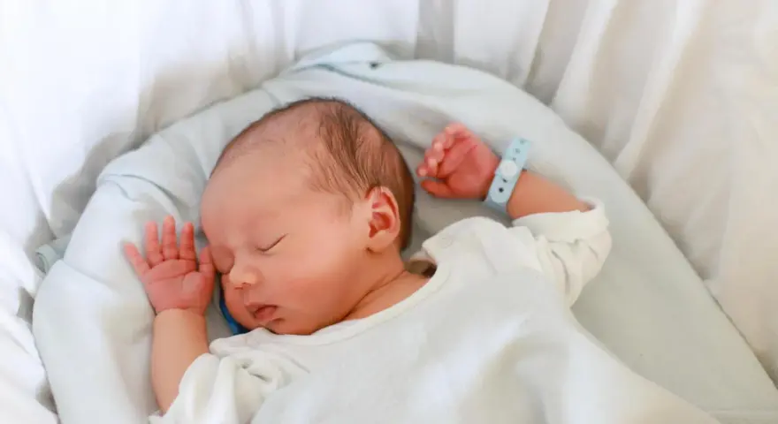 Bebé recién nacido duerme plácidamente en su cuna. Su pulsera de identificación, con el **lua nombre**, indica que está seguro y cuidado.