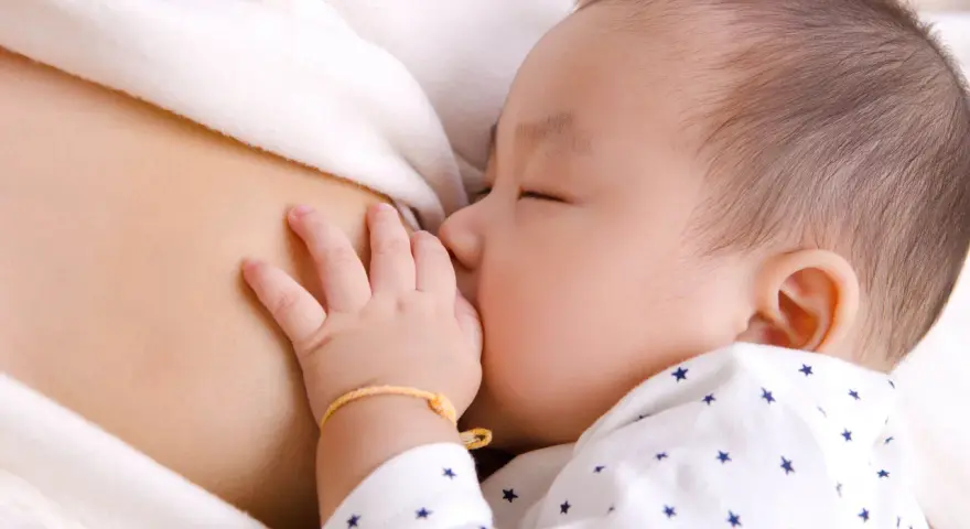 Bebé asiático durmiendo mientras se alimenta, mostrando el reflejo de succión. Su mano pequeña descansa sobre el pecho de su madre.