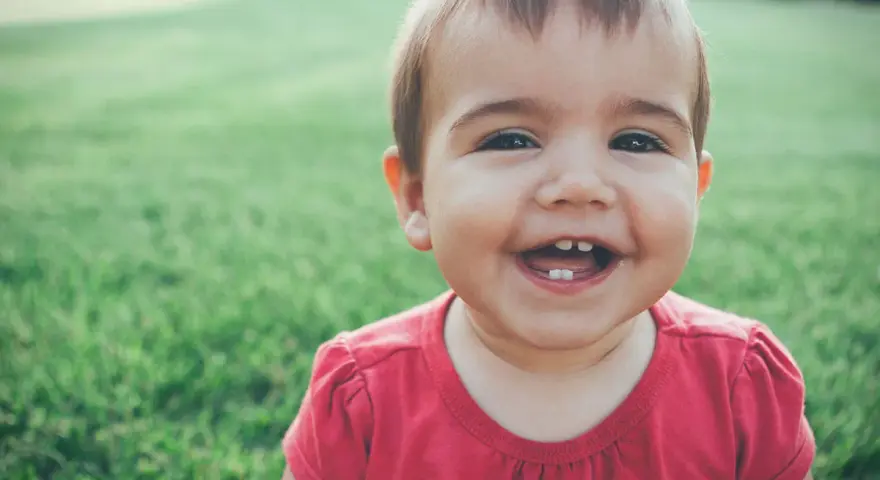 Un bebé de 1 año sin dientes ríe a carcajadas en el césped, mostrando dos pequeños dientes inferiores.