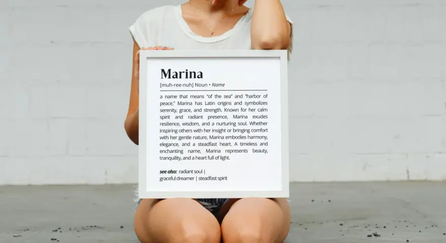 Una mujer sostiene un marco blanco con la definición del nombre Marina, que significa "del mar" y "puerto de paz".