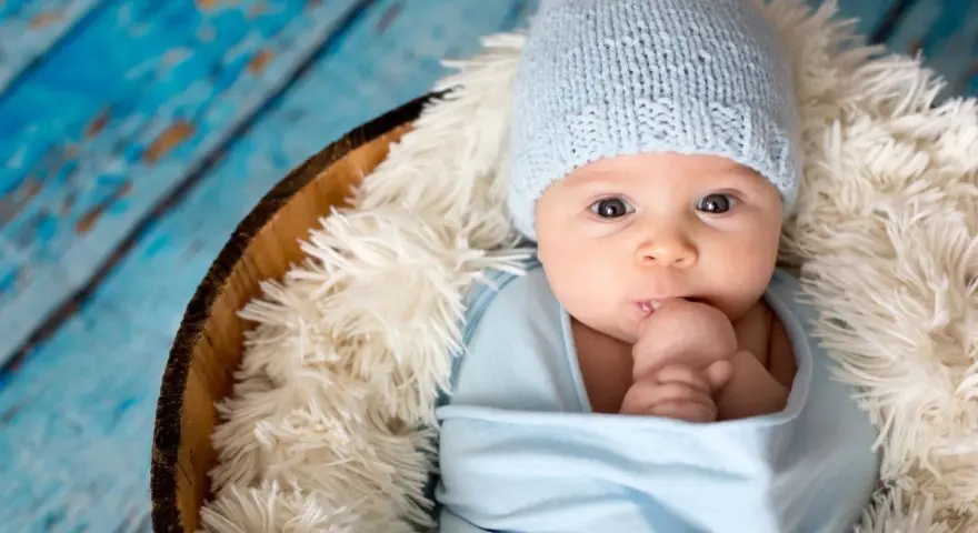 Bebé Ezra, con gorrito azul y envuelto en mantita, mira curioso. Descansa en una cesta sobre fondo de madera azul.