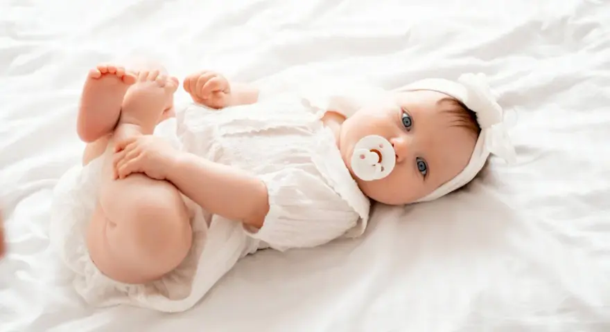 Bebé Ariadna, con su chupete y diadema blanca, juega con sus pies sobre una cama blanca.