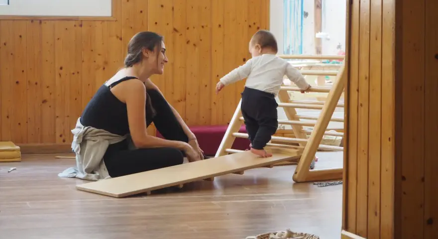 Niña gateando en rampa de madera, animada por mujer sonriente. Fomentando el movimiento libre y la exploración.
