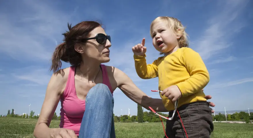 Niña rubia con camiseta amarilla, con el dedo levantado, parece que está aprendiendo cuando empiezan a balbucear los bebes. Madre con gafas de sol la mira.