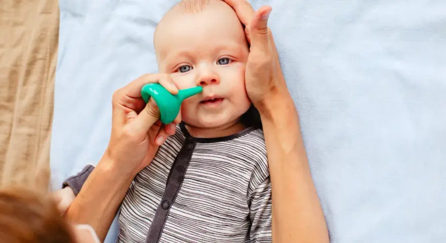 Bebé con ojos azules siendo ayudado a quitar mocos con un aspirador nasal verde.