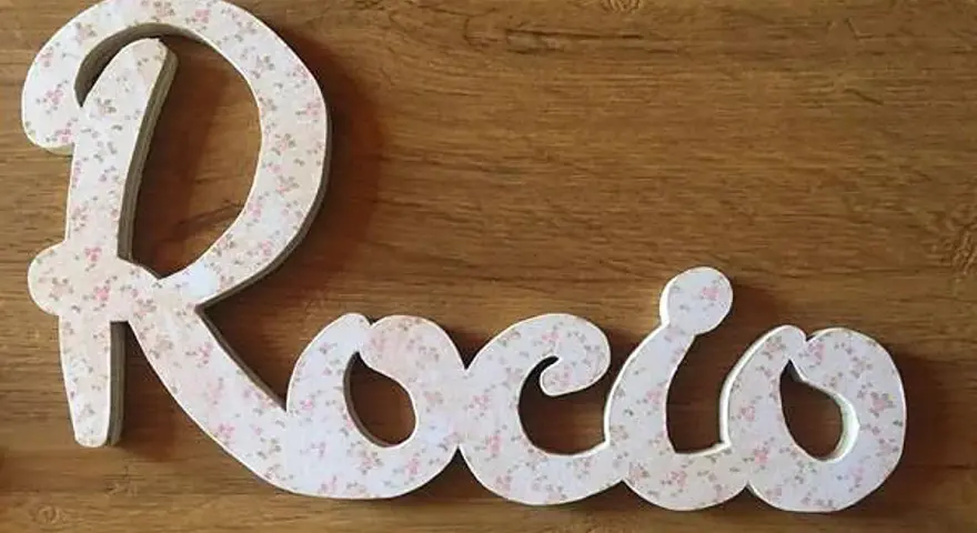 Letra decorativa con el nombre Rocio, cubierta de un delicado estampado floral rosa sobre fondo blanco, sobre una superficie de madera.