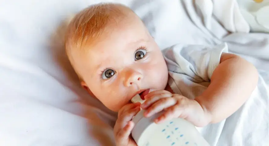 Bebé con ojos grandes y curiosos, bebiendo leche de un biberón. La imagen responde a la pregunta: cuanta leche toma un bebe.