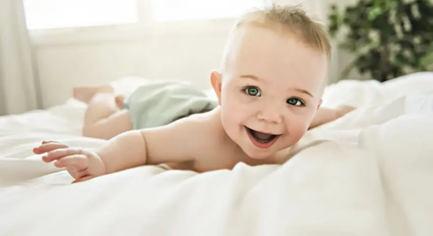 Bebé sonriente boca abajo en una cama blanca, con un reflejo de hociqueo en su expresión juguetona.